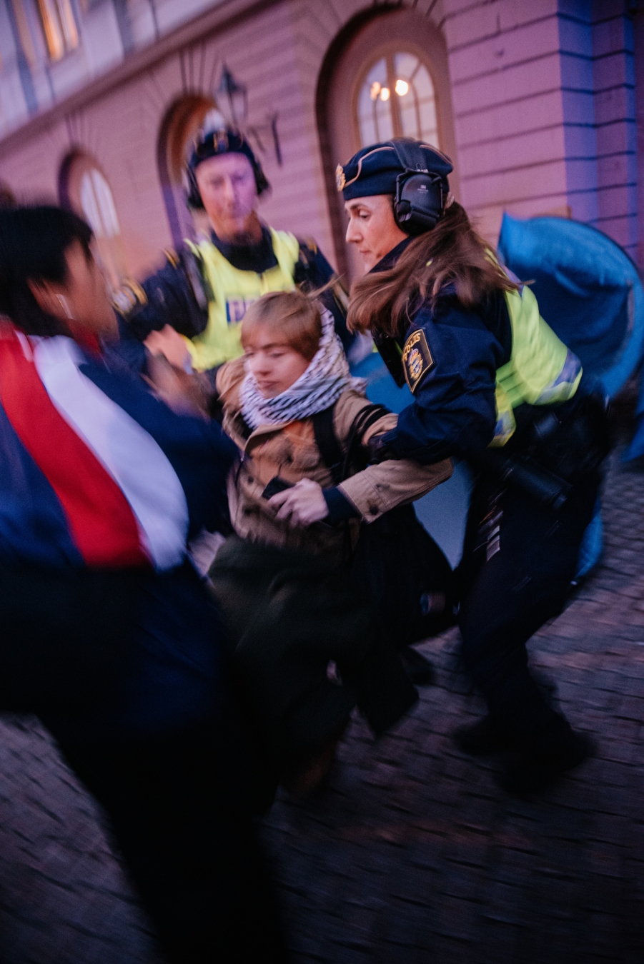 We can see 2 police person is carrying one protester from the tent and third person is asking question. Someone is trying to destroy the blue tents in the behind.